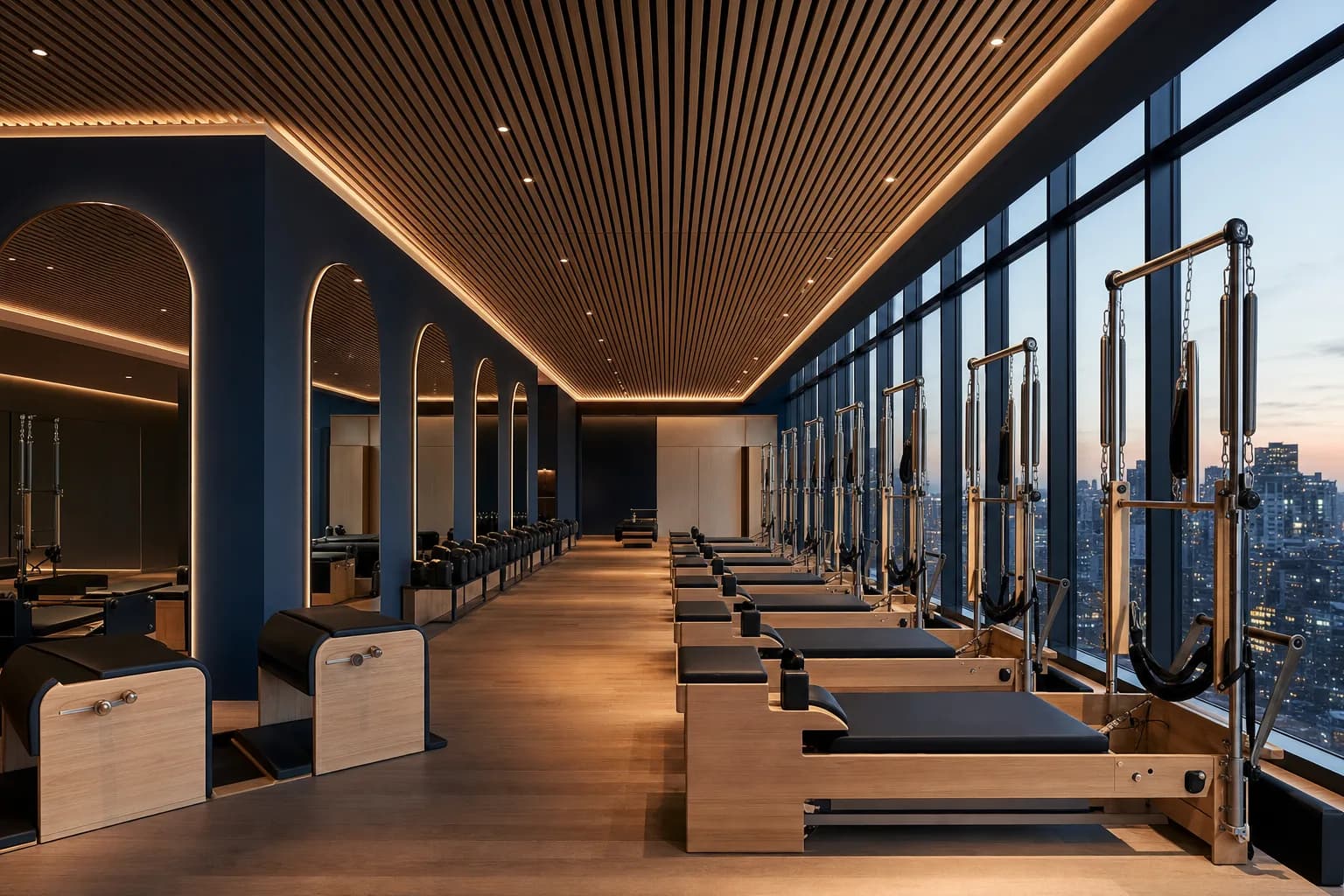 An upscale pilates studio with a row of reformer machines beside floor-to-ceiling windows at dusk